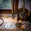An ornate brass and black metal candle holder with a sculpted figure base, holding two tall candle sleeves, displayed on a tabletop next to a stained-glass panel and a wooden clock.