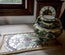 A decorative ceramic pitcher with green, yellow, and black floral patterns sits on a matching embroidered runner in front of a wooden clock and window.