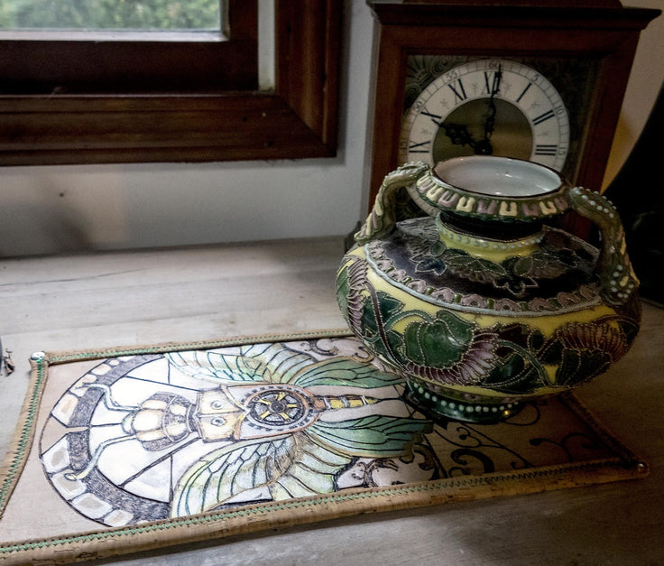 A decorative ceramic pitcher with green, yellow, and black floral patterns sits on a matching embroidered runner in front of a wooden clock and window.