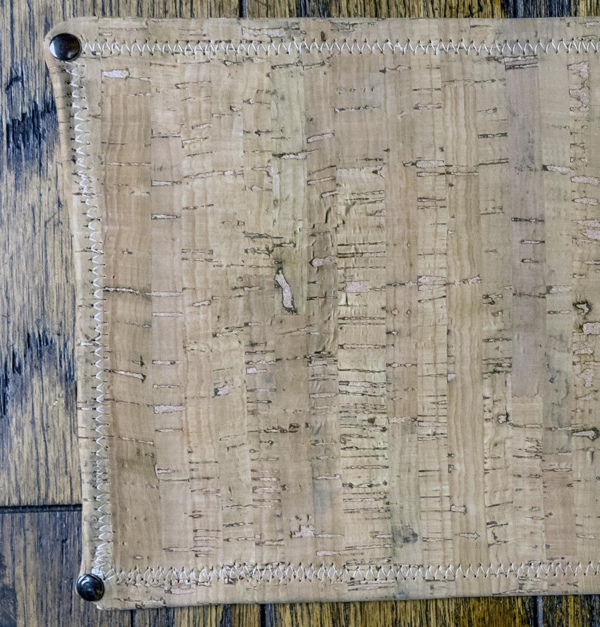 A close-up of a rectangular cork mat with stitched edges and metal corner protectors, laid flat on a dark wood surface.