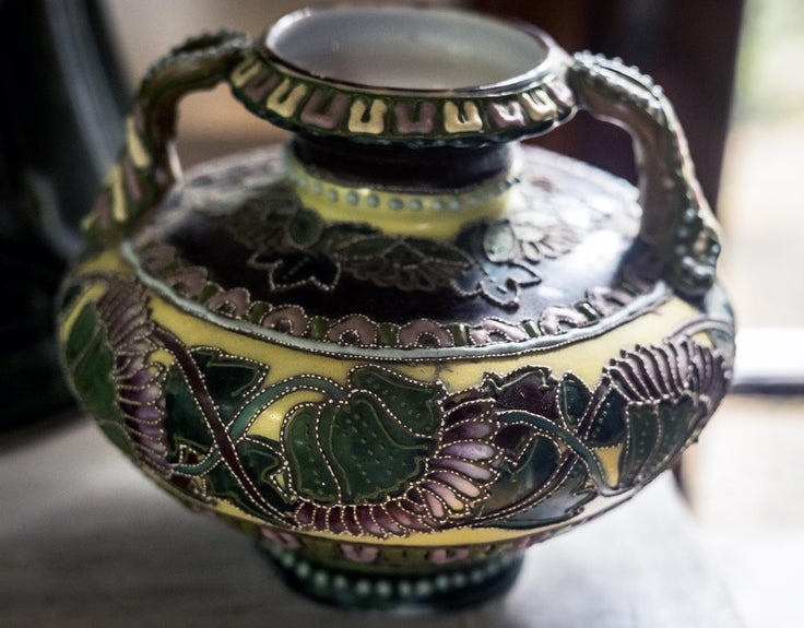 An ornate vintage ceramic vase with a dark brown and yellow floral pattern, featuring green and purple enamel details and decorative handles.